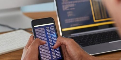 Unrecognizable African-American man holding smartphone with code on screen while working at desk in office, IT developer concept, copy space